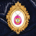 Art Fairy Magic pendant framed in ornate gold on a dark fabric background, with a pink oval charm