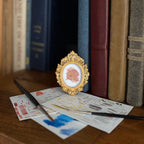 Art and Magic theme fairy illustration in a gold frame beside stacked books and papers on a wooden table