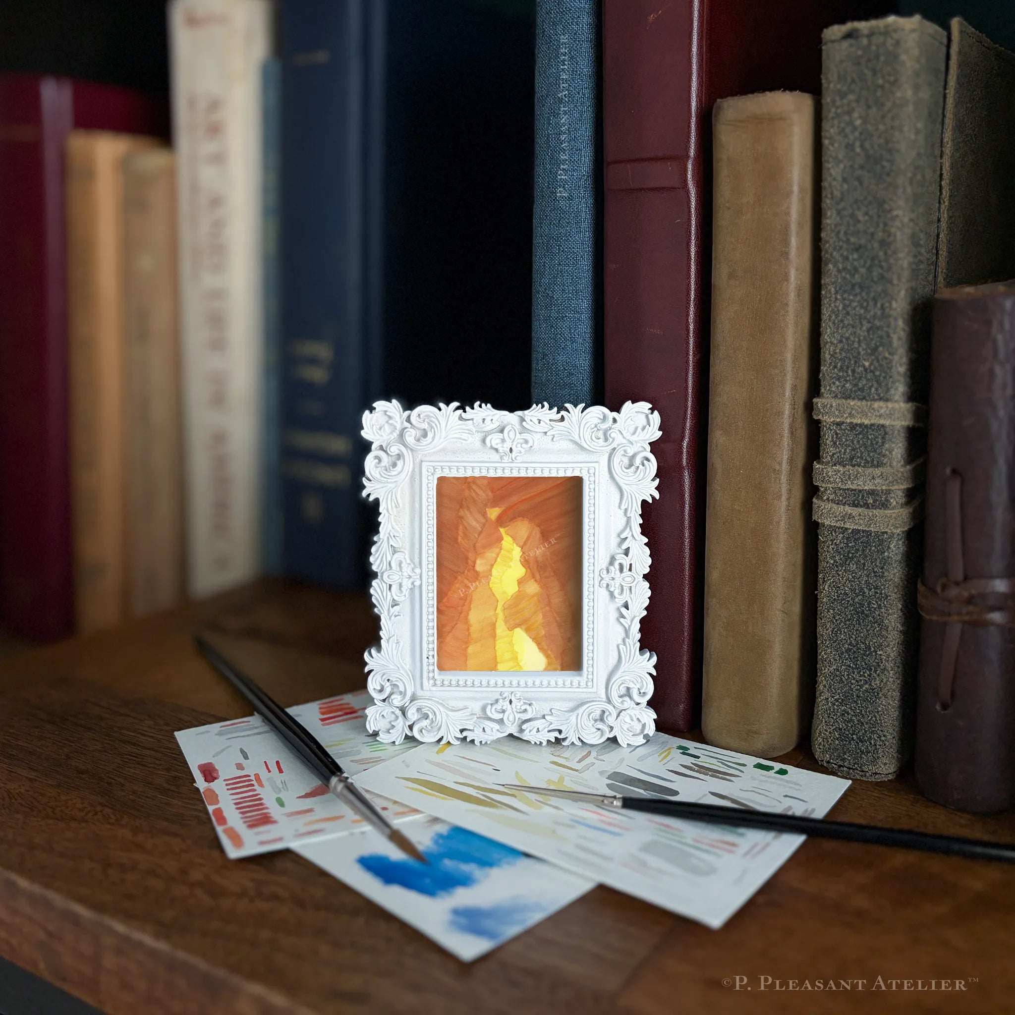 A small white ornate picture frame holding a glowing orange candle, styled on a wooden table in front of a bookshelf with books and paintbrushes.