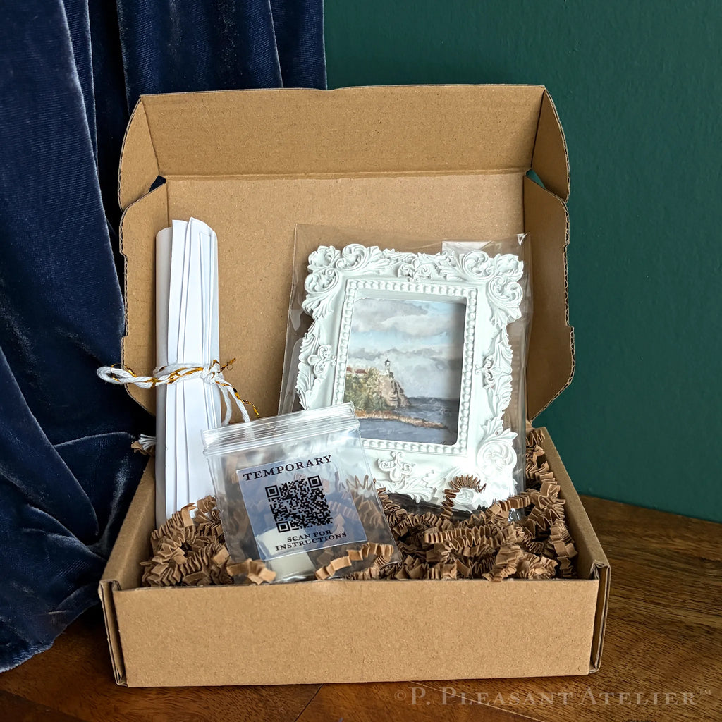 A curated gift box from P. Pleasant Atelier containing a small framed landscape print in an ornate white frame, a bag of installation putty for temporary installation, rolled white papers, all nestled in brown crinkle paper inside a brown cardboard box.