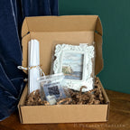 A curated gift box from P. Pleasant Atelier containing a small framed landscape print in an ornate white frame, a bag of installation putty for temporary installation, rolled white papers, all nestled in brown crinkle paper inside a brown cardboard box.