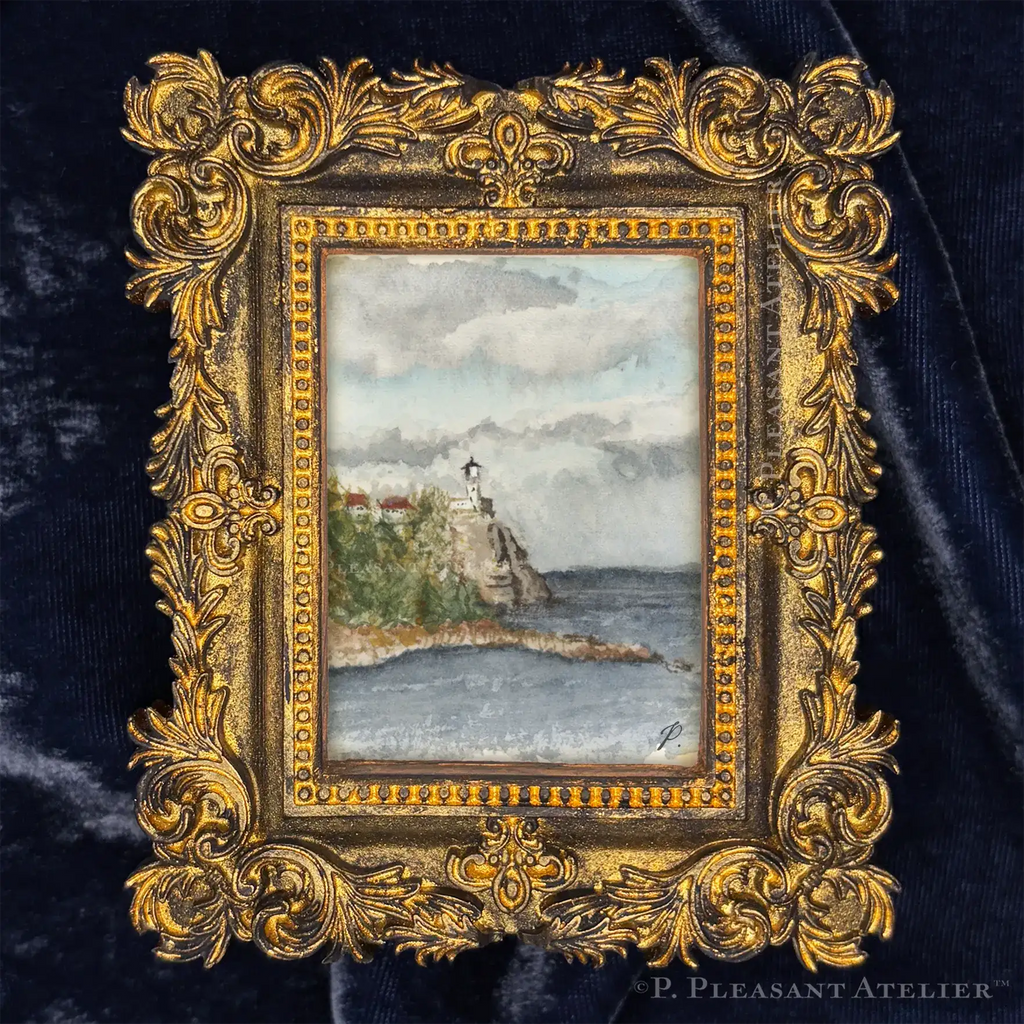 A miniature framed art piece featuring a hand-painted lighthouse scene on a rocky coast, set within an ornate gold frame and displayed on a dark navy velvet background.