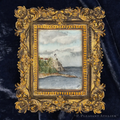 A miniature framed art piece featuring a hand-painted lighthouse scene on a rocky coast, set within an ornate gold frame and displayed on a dark navy velvet background.