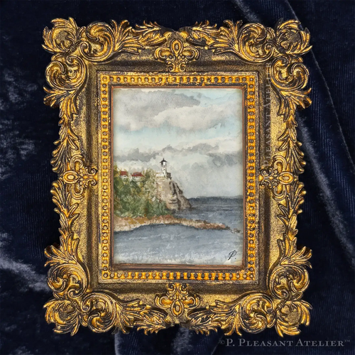 A miniature framed art piece featuring a hand-painted lighthouse scene on a rocky coast, set within an ornate gold frame and displayed on a dark navy velvet background.