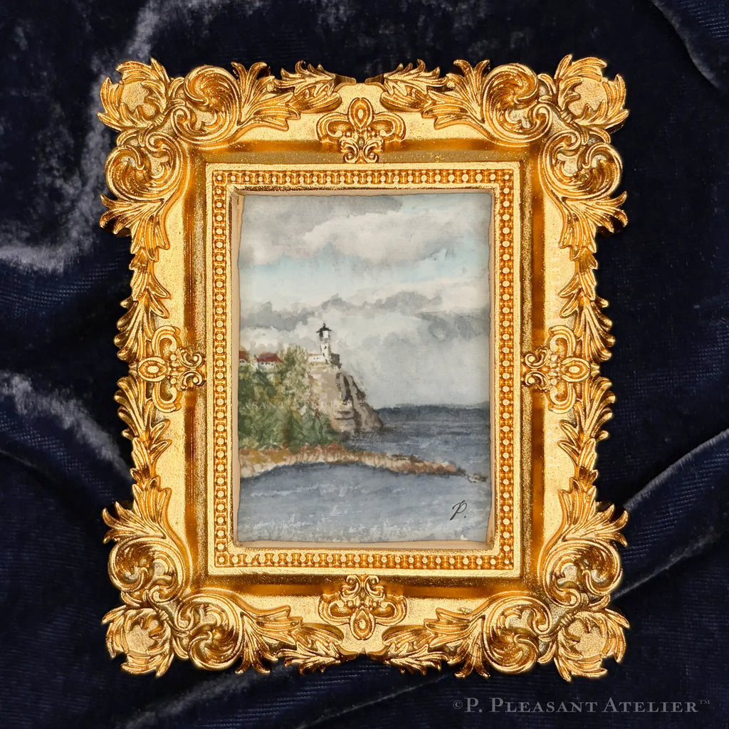 A small framed watercolor painting of a lighthouse by the sea, set in an ornate gold frame against a dark blue velvet background.