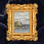 A small framed watercolor painting of a lighthouse by the sea, set in an ornate gold frame against a dark blue velvet background.