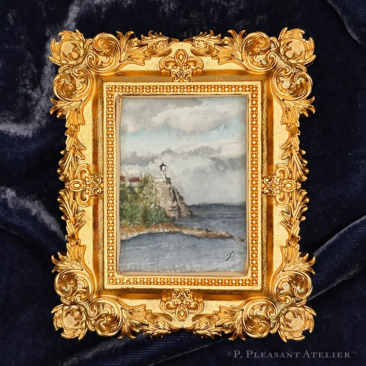 A small framed watercolor painting of a lighthouse by the sea, set in an ornate gold frame against a dark blue velvet background.