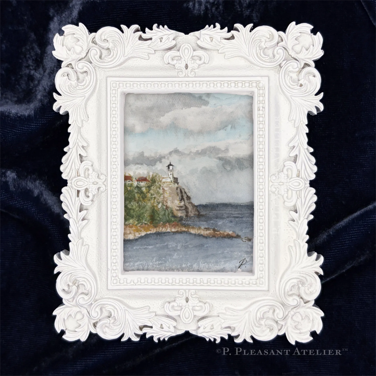 An ornate white baroque-style picture frame displaying a watercolor painting Split Rock lighthouse on a rocky cliff by Lake Superior, set against a dark navy blue velvet background. Signed by P. Pleasant with a "P."