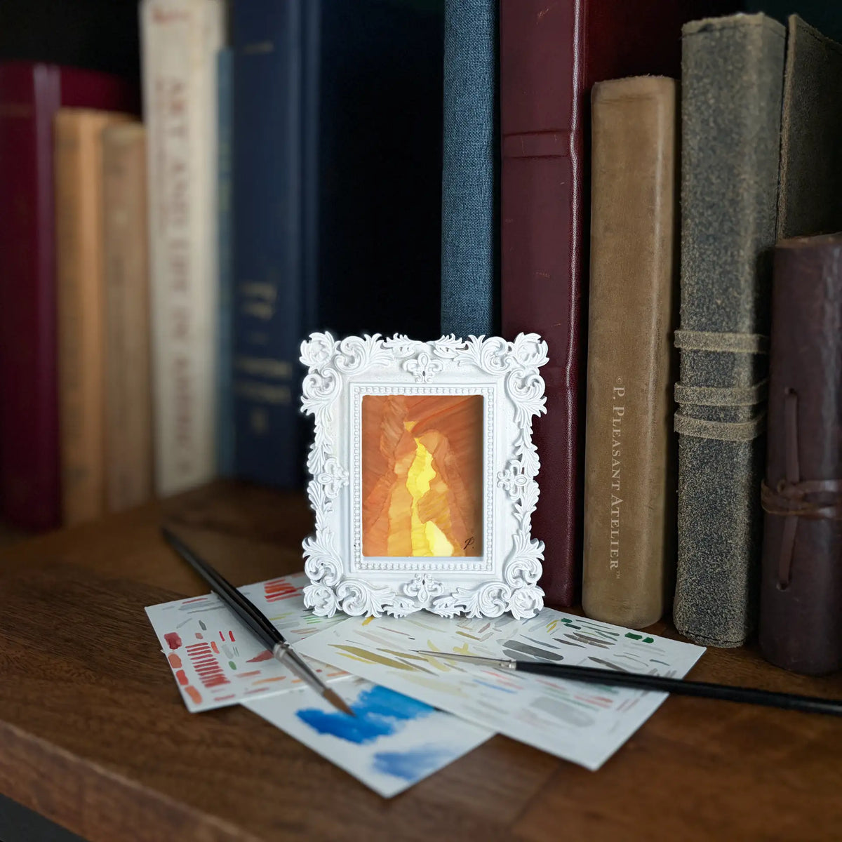 A small white ornate picture frame holding a painting from inside a desert canyon, styled on a wooden table in front of a bookshelf with books and paintbrushes.