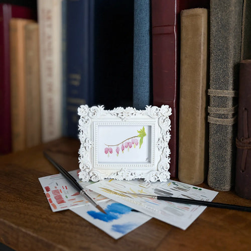A small white ornate picture frame holding a watercolor illustration of pink bell-shaped flowers, styled on a wooden table with paintbrushes, paint swatches, and books in the background.