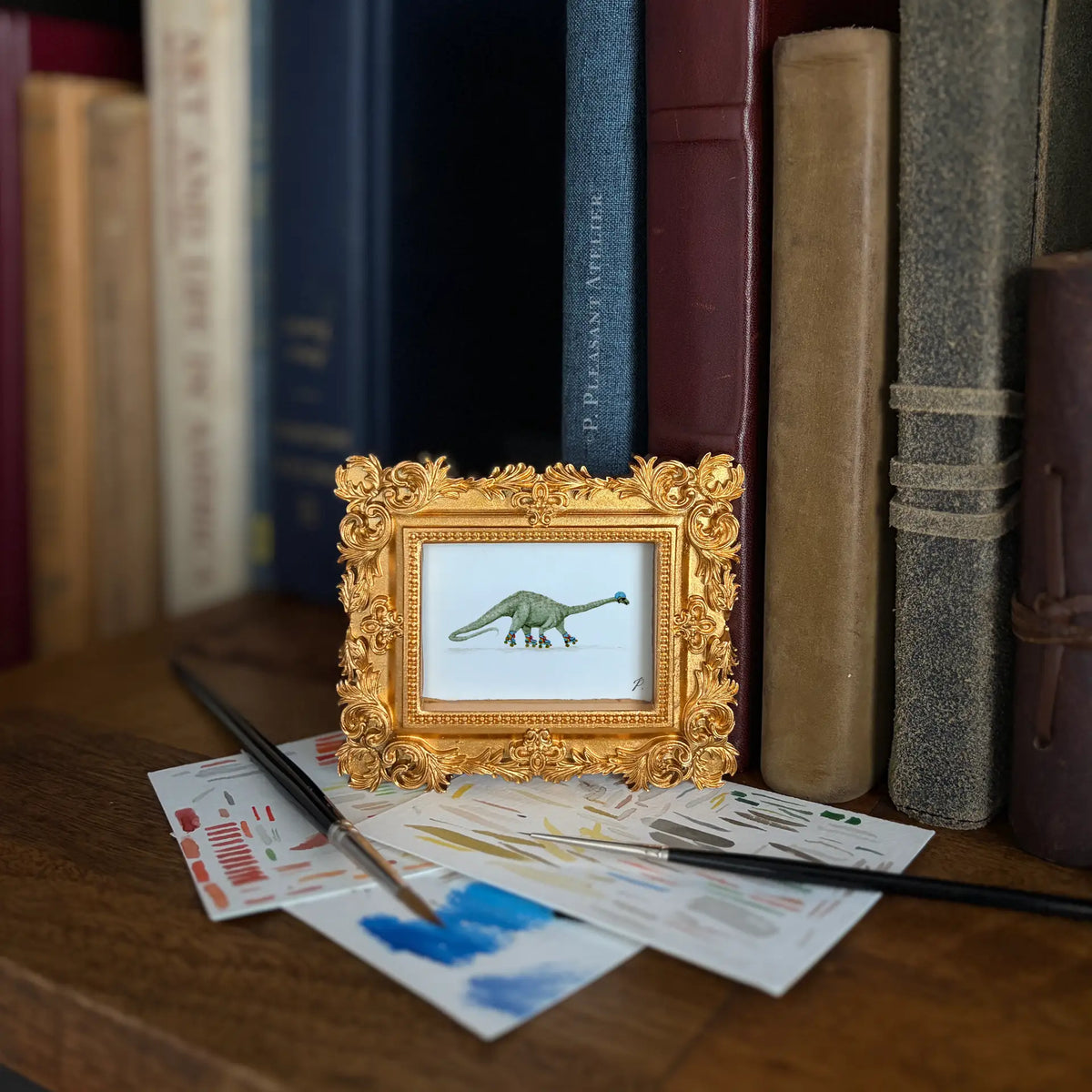 A small framed dinosaur art print with an ornate gold frame sits on a wooden table in front of a row of books. Paintbrushes and paint swatches are scattered nearby.