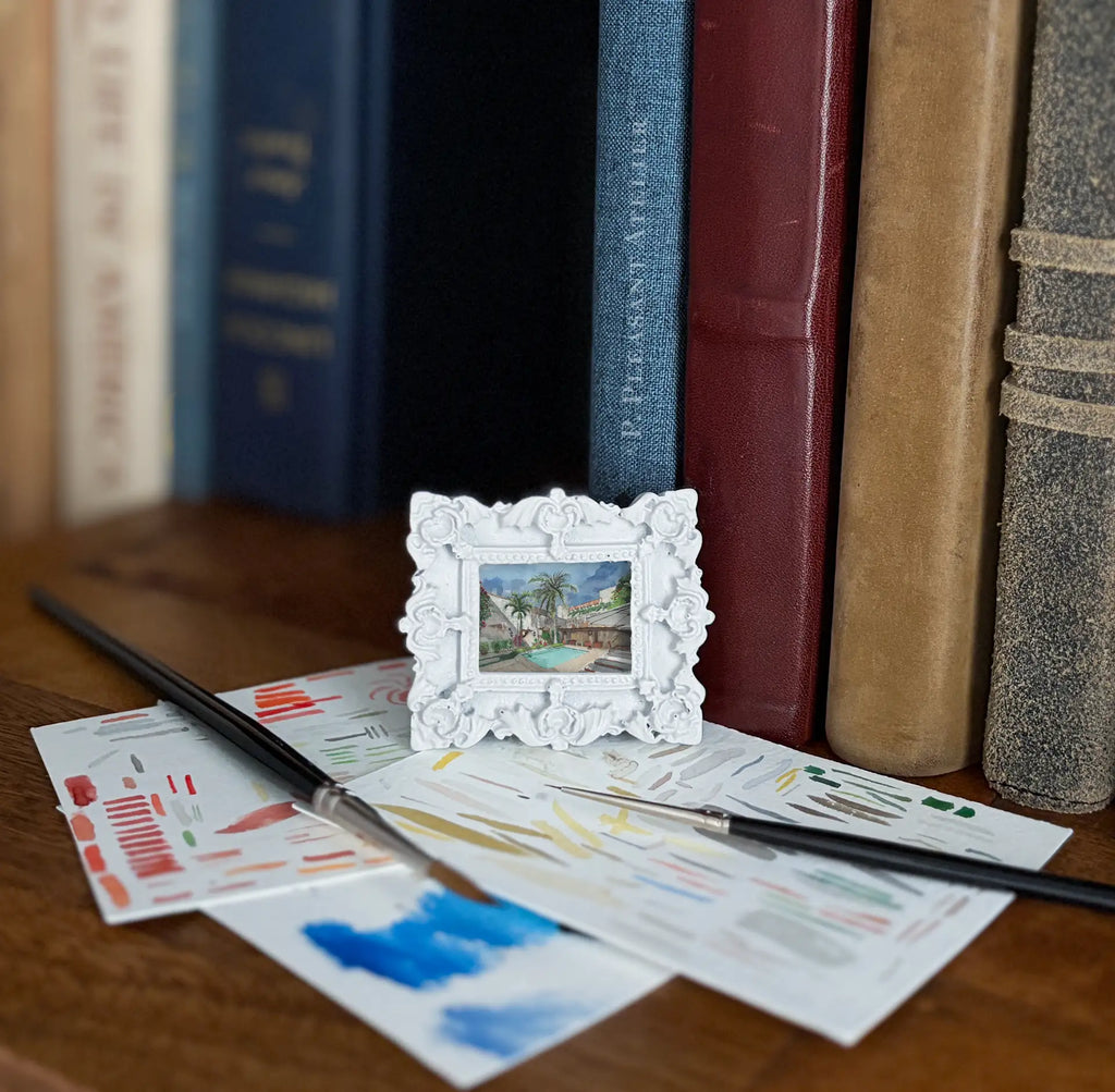 A miniature white ornate picture frame holding a small landscape painting, styled on a wooden surface with paint swatches, paintbrushes, and books in the background.