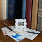 A miniature white ornate picture frame holding a small landscape painting, styled on a wooden surface with paint swatches, paintbrushes, and books in the background.