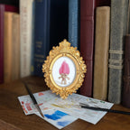 A small ornate gold-framed art print featuring a pink-haired character, styled on a wooden table in front of a bookshelf with books and paintbrushes.