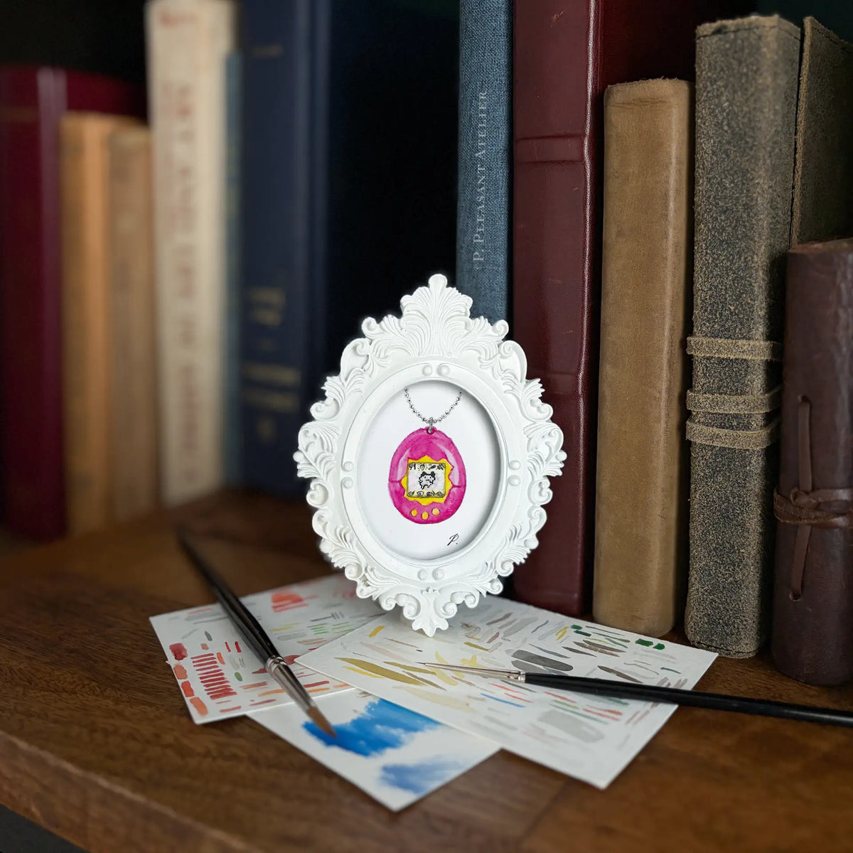 A small white ornate oval frame holding a painting of a pink and colorcul circular charm, displayed on a wooden shelf in front of vintage books with paintbrushes and paint cards nearby.