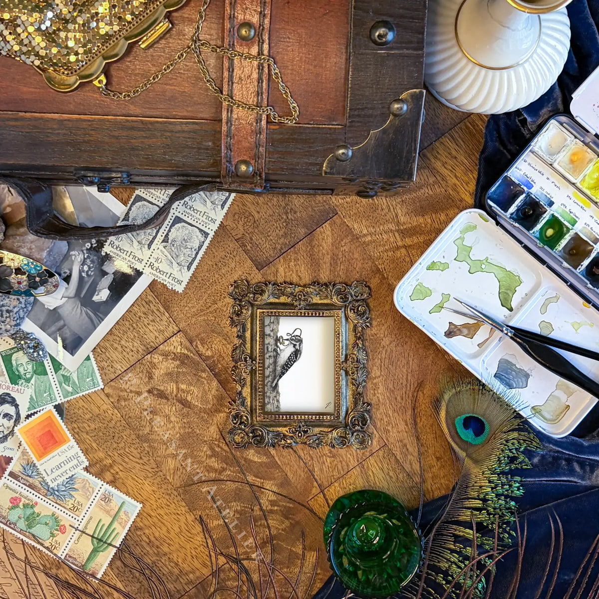 A styled flat-lay of a small framed art print featuring a black bird illustration on a white background, surrounded by vintage postage stamps, a wooden chest, a paint palette, and a peacock feather on a wooden surface.