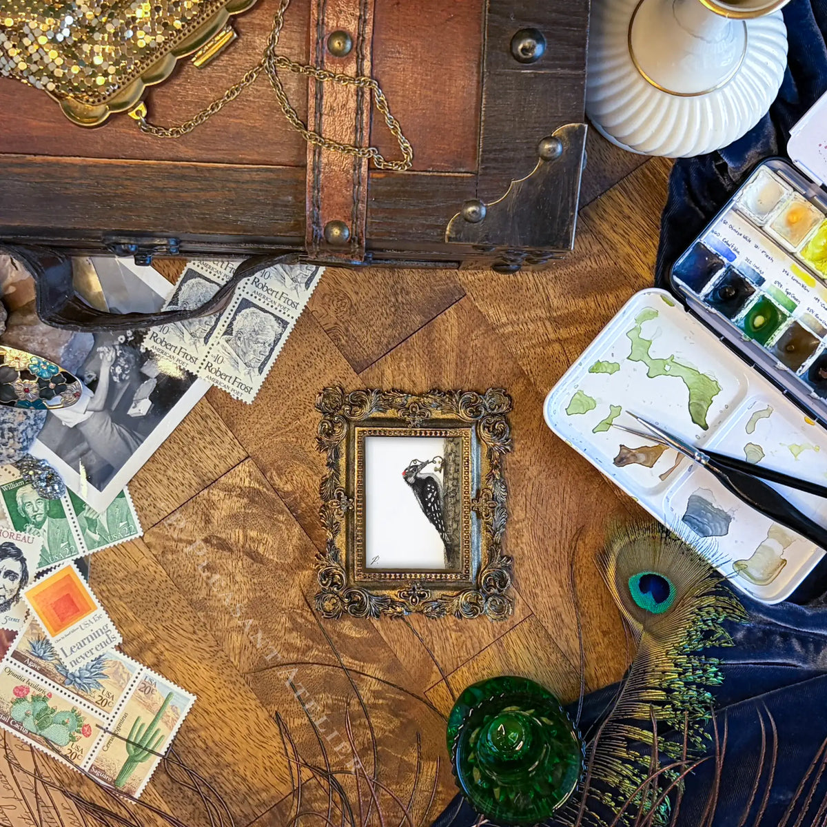 A styled flat-lay featuring a small, ornate gold picture frame containing a black and white bird illustration, surrounded by vintage postage stamps, a wooden chest, a watercolor palette, and a peacock feather on a rustic wooden table.