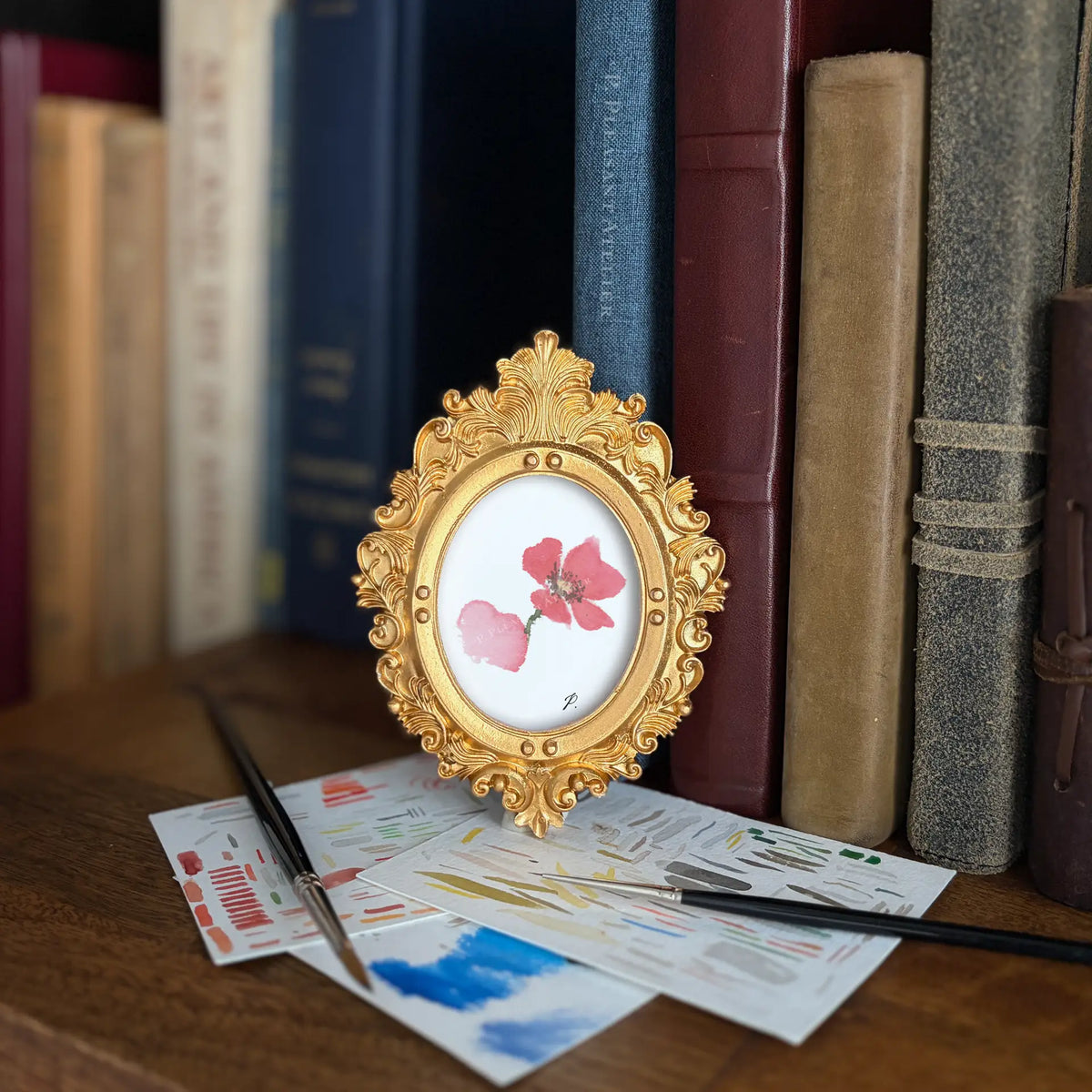 An ornate gold oval picture frame holding a pressed pink flower, styled on a wooden surface with paintbrushes and art papers in front, and a blurred row of books in the background.