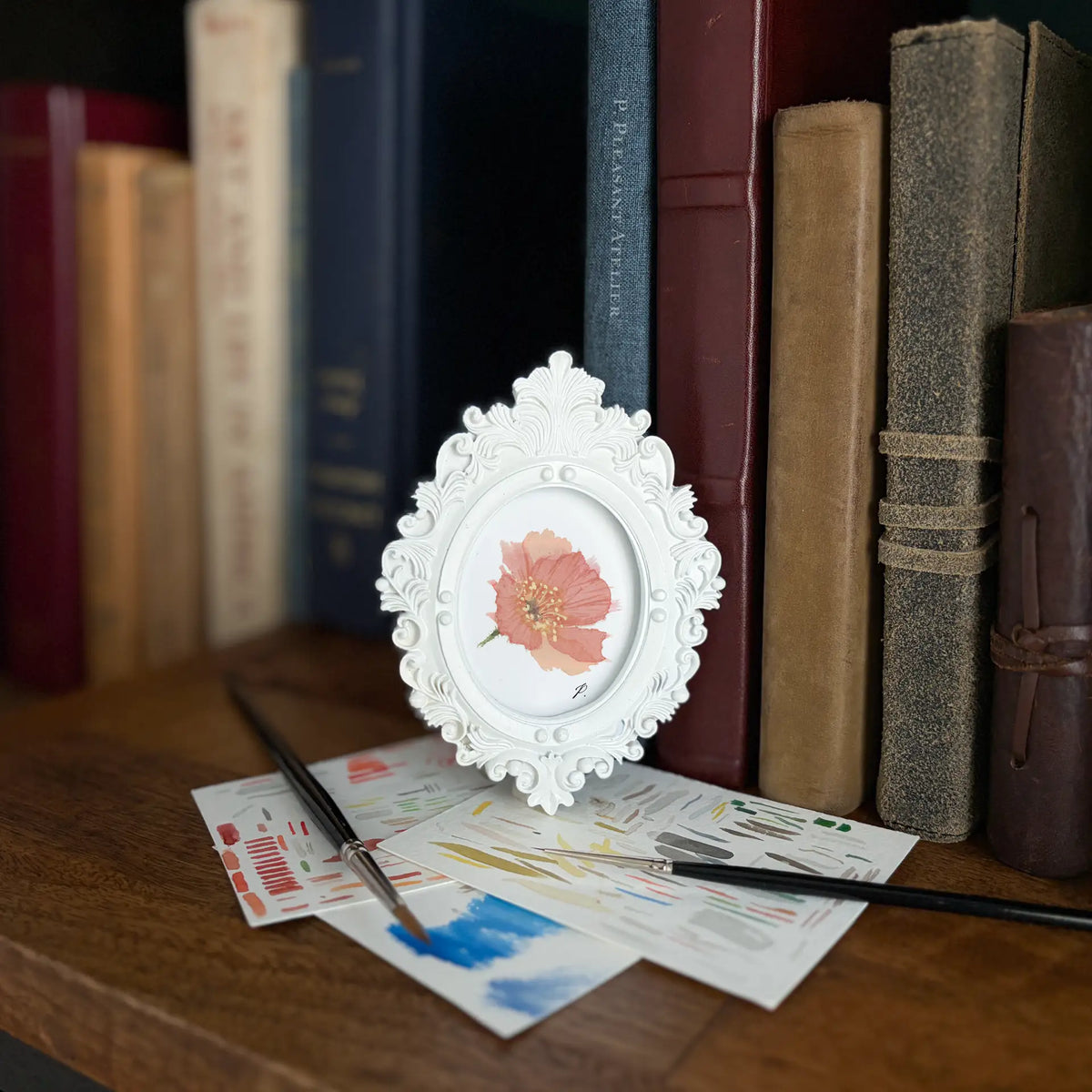 A small white ornate oval picture frame holding a pink flower illustration, styled on a wooden table with paintbrushes and art papers in front of a background of book spines.