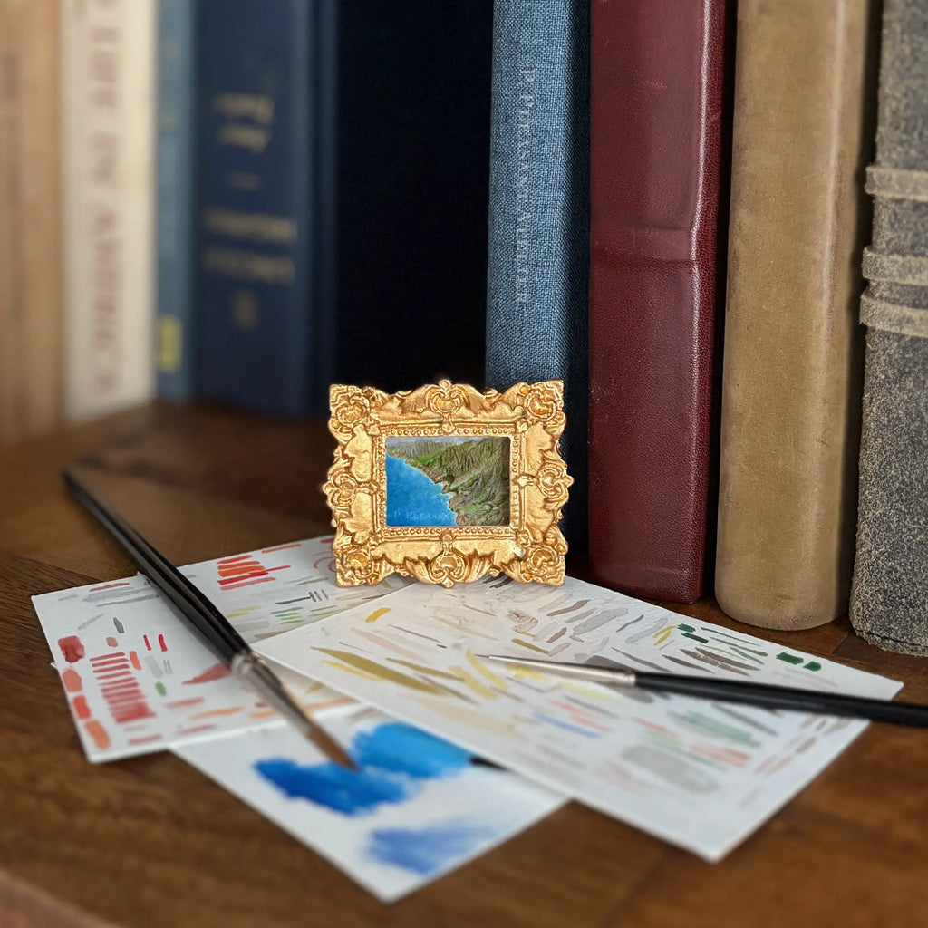 A small ornate gold picture frame holding a landscape painting of a blue ocean and green cliffs, styled on a wooden table with paintbrushes, paint swatches, and books in the background.