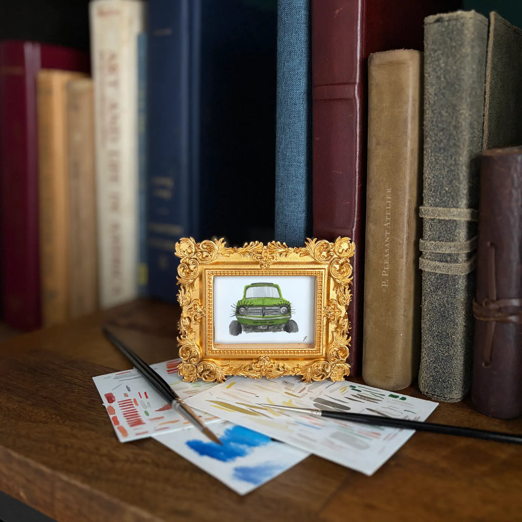 Art fairy magic framed illustration of a green car in ornate gold small frame. Styled on wooden shelf with a background of books and close to watercolor tests with paintbrushes.