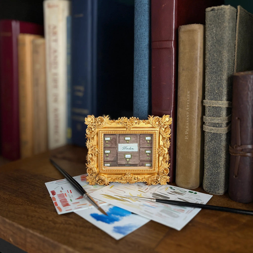 A miniature gold-framed picture frame with a floral baroque border, holding a painting of a card catalogue with "Windom" written on a card coming out of a drawer. The frame sits on a wooden shelf surrounded by old books, paintbrushes, and paint swatches.