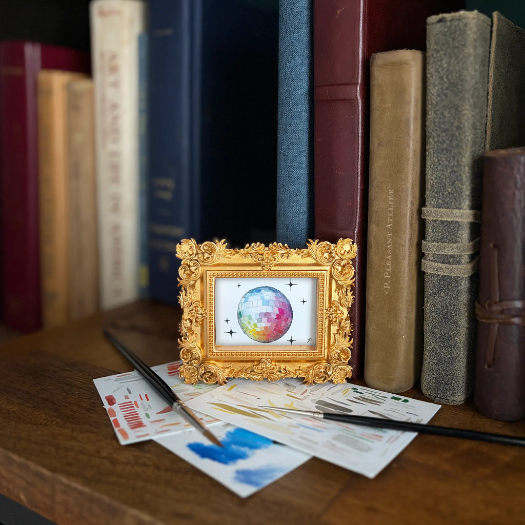 A small ornate gold picture frame holding a colorful painting of a disco ball, styled on a wooden shelf in front of books with paintbrushes and paint swatches nearby.