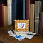 A small ornate gold picture frame holding a colorful painting of a disco ball, styled on a wooden shelf in front of books with paintbrushes and paint swatches nearby.