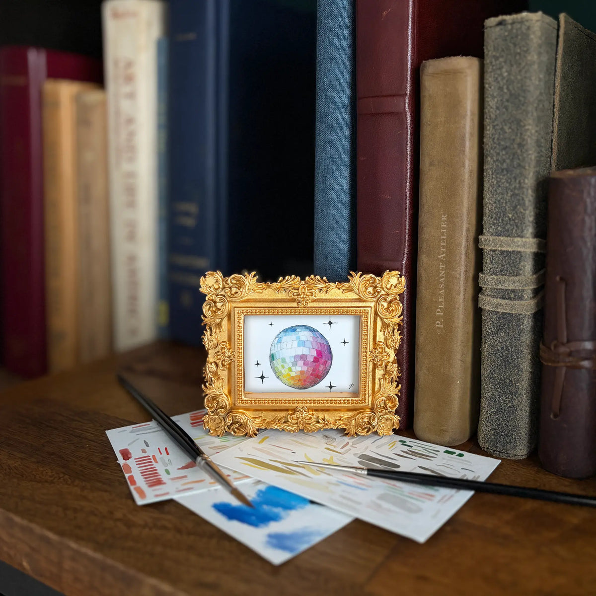 A small ornate gold picture frame holding a colorful painting of a disco ball, styled on a wooden shelf in front of books with paintbrushes and paint swatches nearby.
