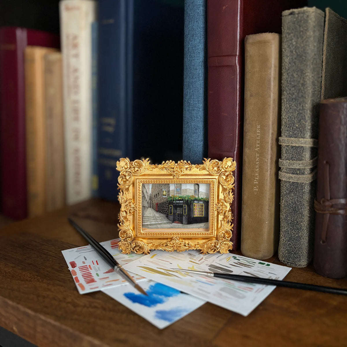A small ornate gold picture frame displaying a painting of a European pub, styled on a wooden shelf in front of vintage books with paintbrushes and paint swatches.