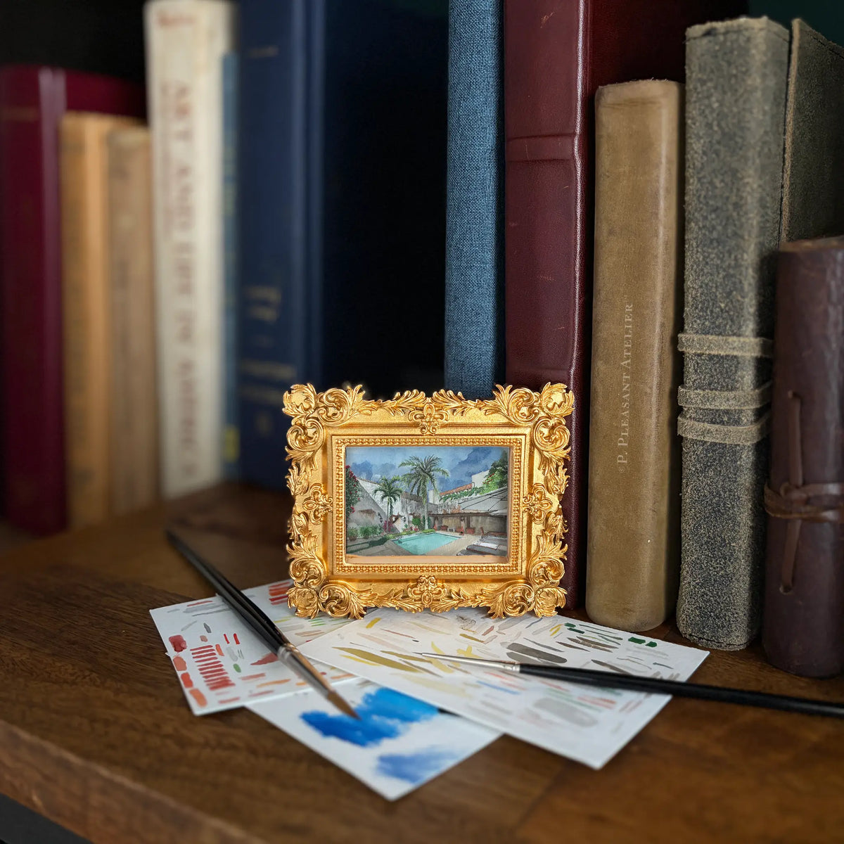 A small ornate gold picture frame holding a colorful painting of a tropical villa and pool, styled on a wooden shelf in front of vintage books with paintbrushes and paint swatches.