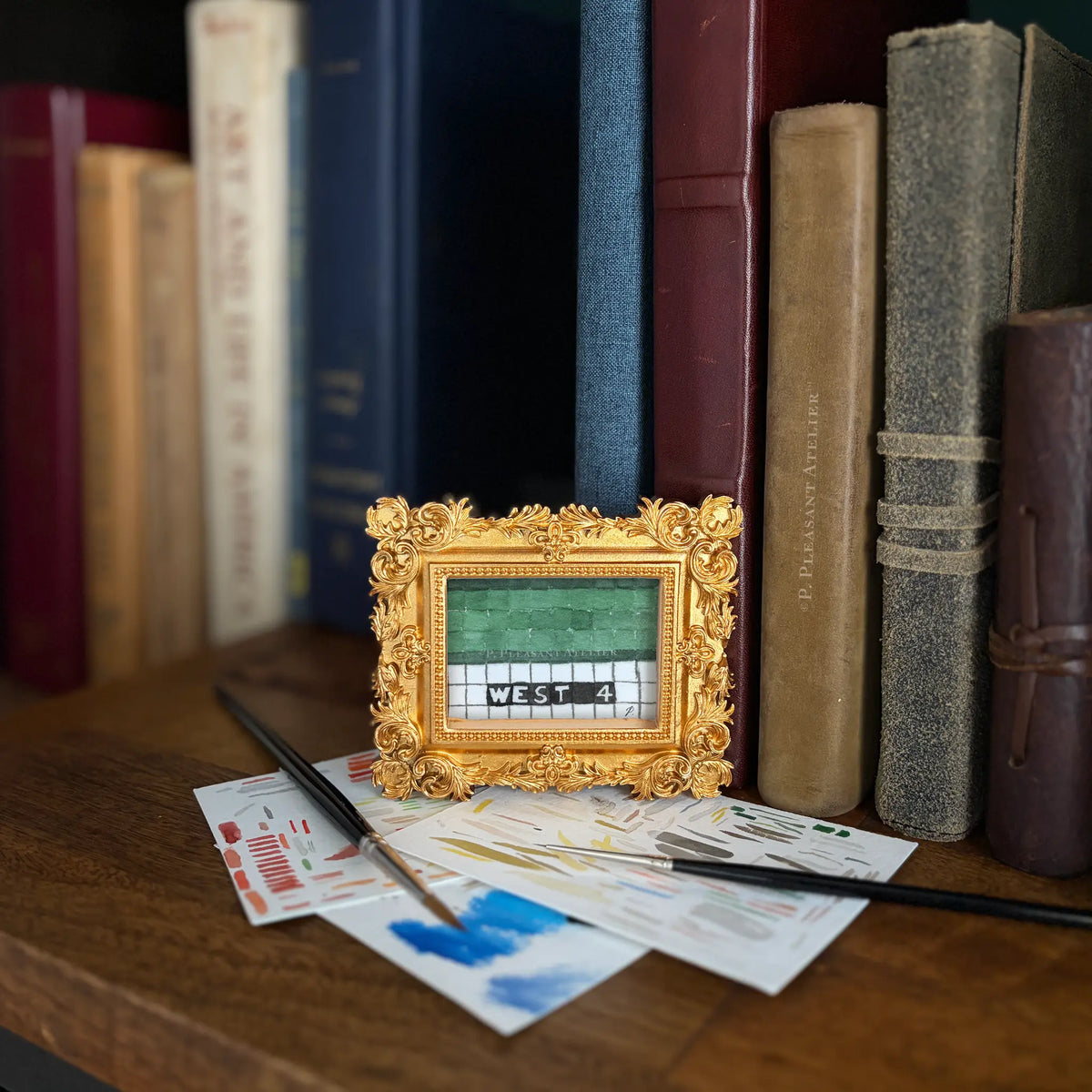 A small ornate gold picture frame holding a painting of subway tiles with letters that read 'WEST 4'. It sits on a wooden table surrounded by paintbrushes, paint swatches, and a row of vintage books in the background.