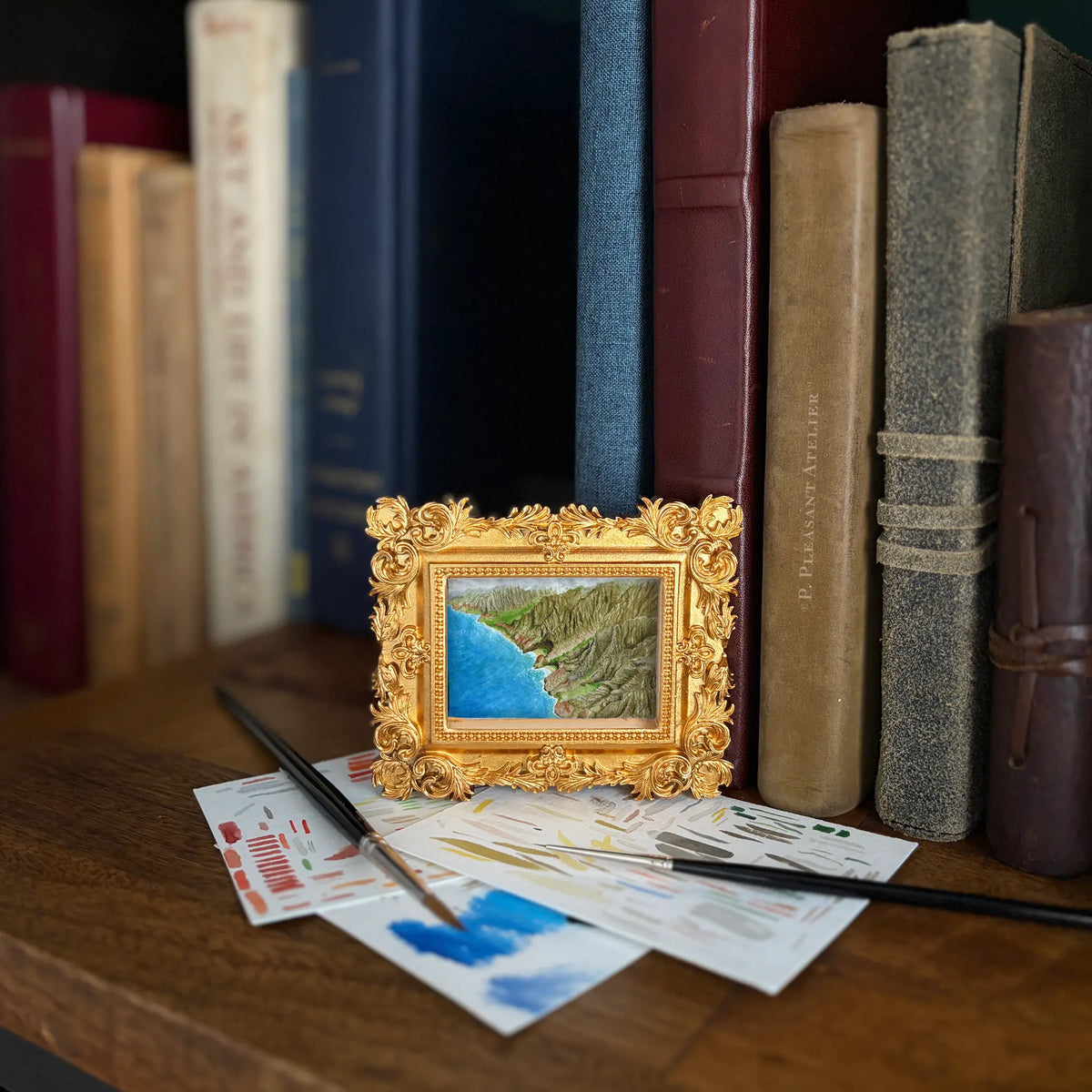 A miniature gold-framed picture of a coastline with blue water, styled on a wooden surface with paintbrushes and paint swatches in front of a row of vintage books.