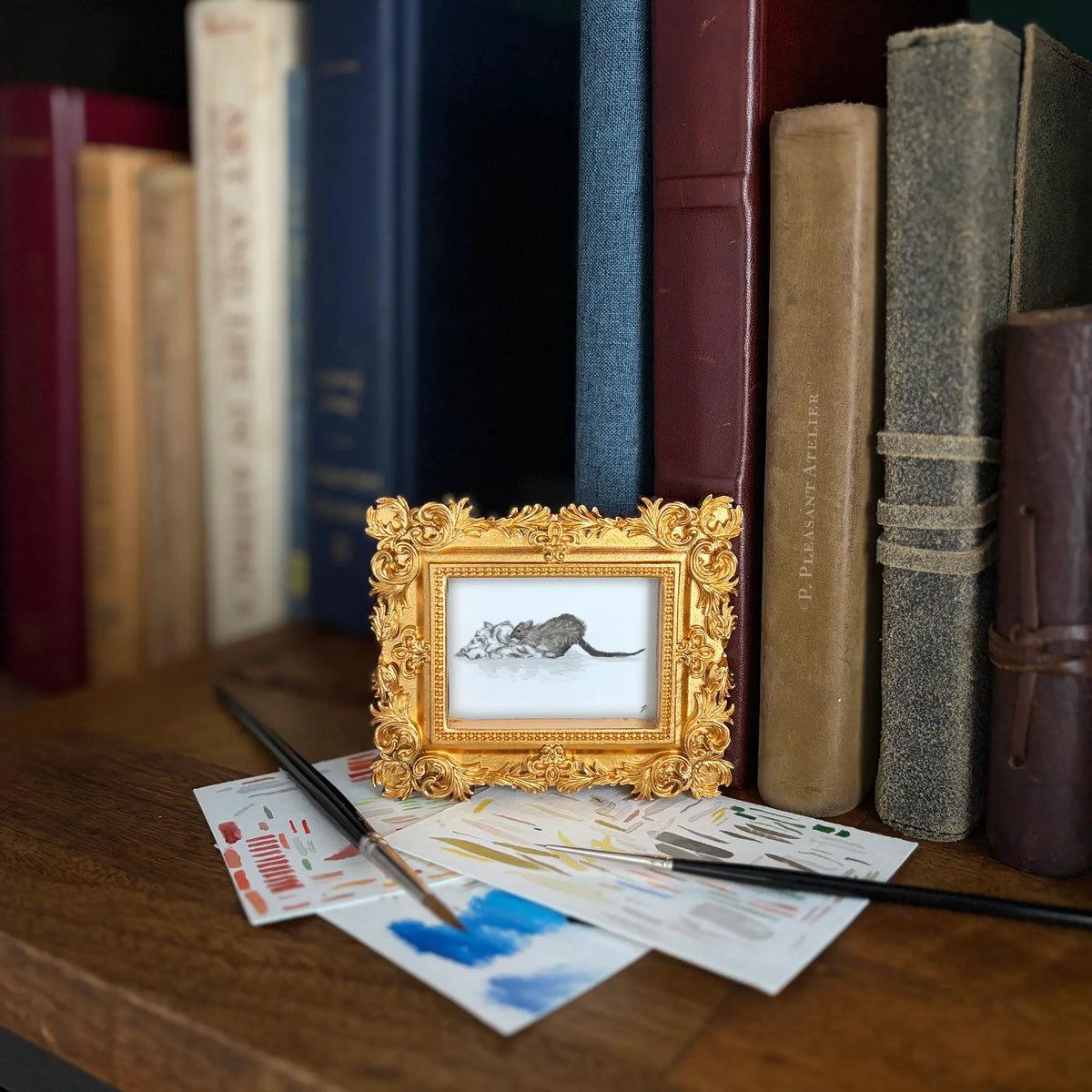 A small ornate gold picture frame holding a painting of a rat on a white background, styled on a wooden surface in front of vintage books with paintbrushes and paint swatches.