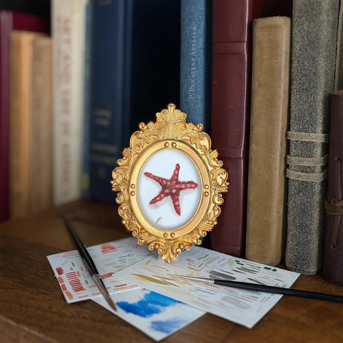 A small ornate gold-framed art print featuring a red starfish on a white background, styled on a wooden surface with paintbrushes and art papers in front, and a row of books in the blurred background.