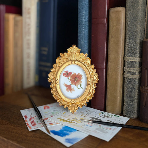 A small oval gold-framed artwork holding a painting of three red and orange poppies, styled on a wooden surface with paintbrushes and paint cards in front, and a row of books in the background.