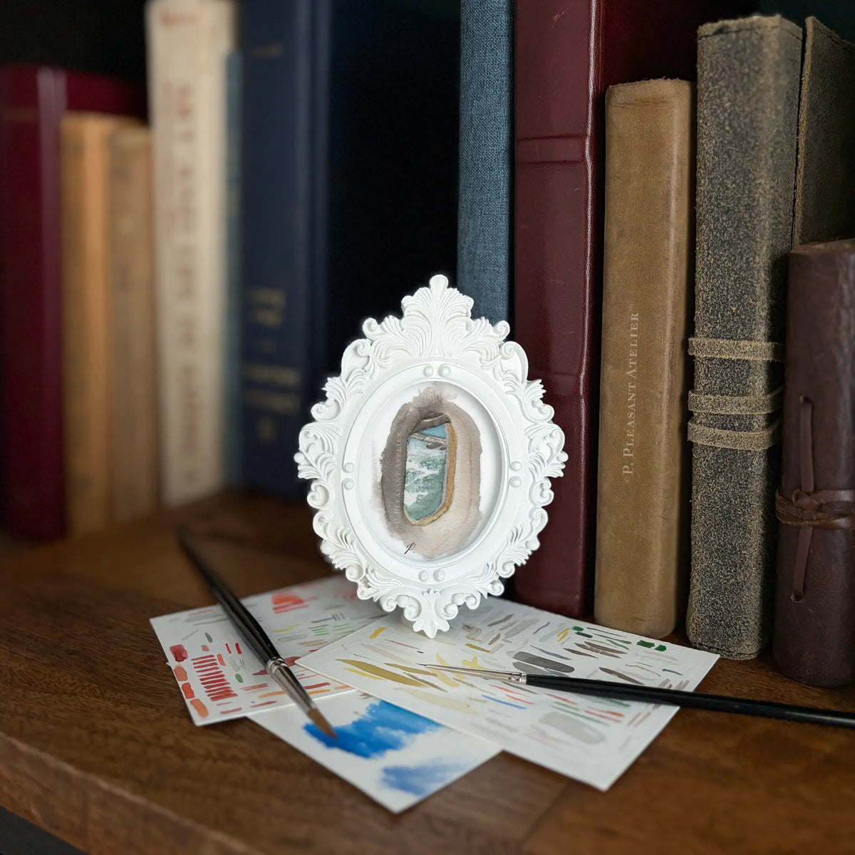 An ornate white frame holding a painting looking out a plane window onto clouds and sky. It sits on a wooden table, surrounded by paintbrushes, paint swatches, and a row of vintage books in the background.