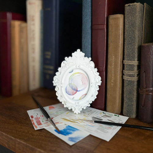 A small white ornate oval picture frame with a colorful painting of bubbles styled on a wooden table next to paintbrushes and art papers, with vintage books in the background.