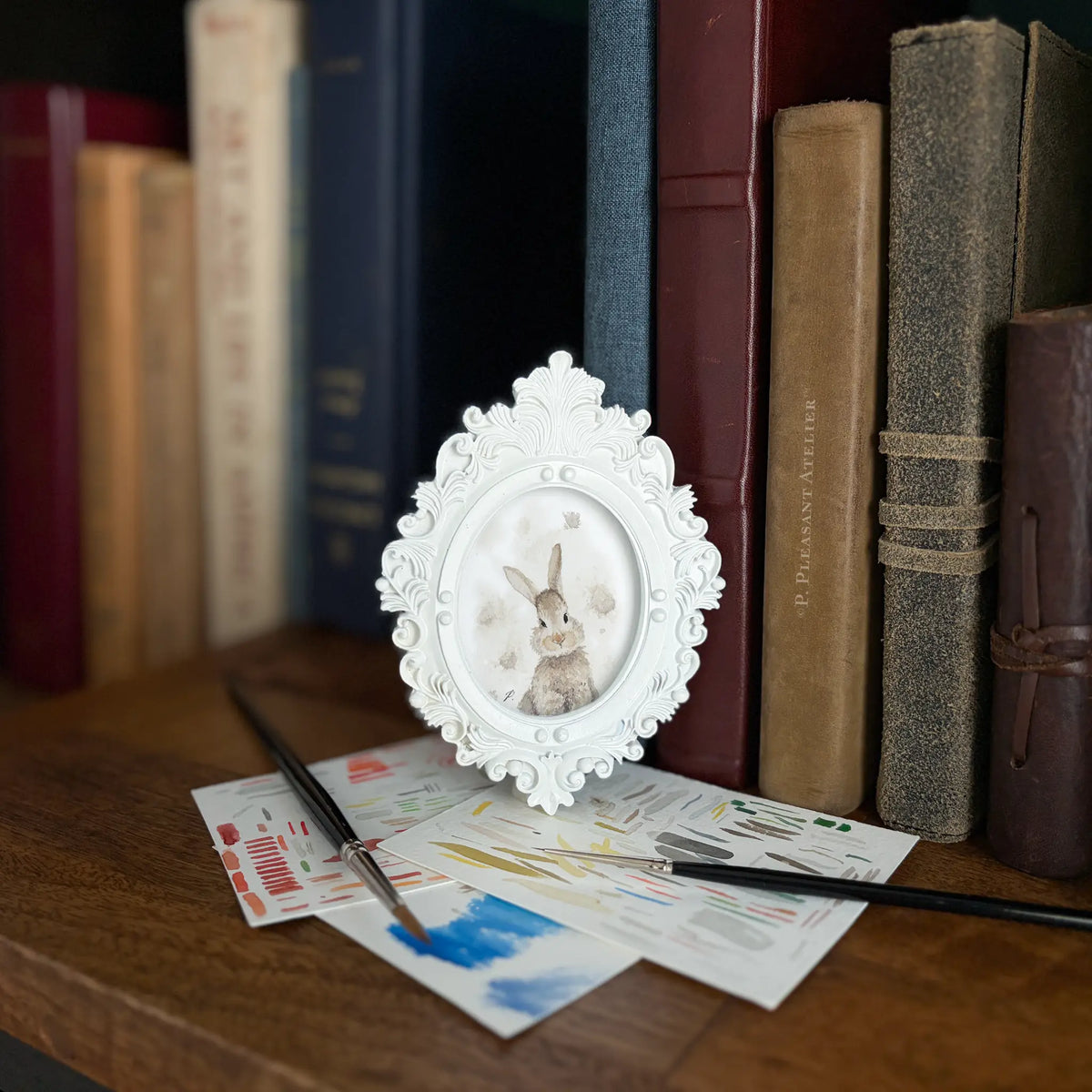 A small white ornate oval picture frame holding a painting of a bunny with a spotted grey and white background, styled on a wooden table with paintbrushes and art papers in front of a background of book spines.
