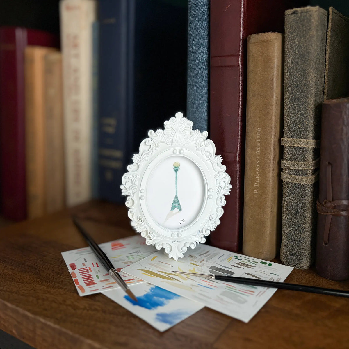 A small white ornate picture frame with a painting of a green lamp post, displayed on a wooden shelf next to books and art supplies.