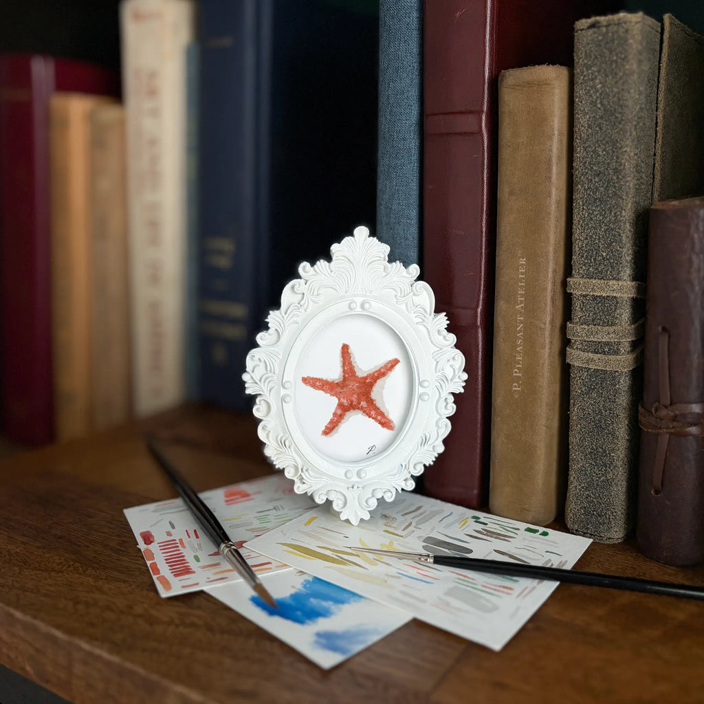 A small white ornate oval picture frame holding painting of a starfish on a white background, styled on a wooden table with paintbrushes and art papers in front of a background of book spines.