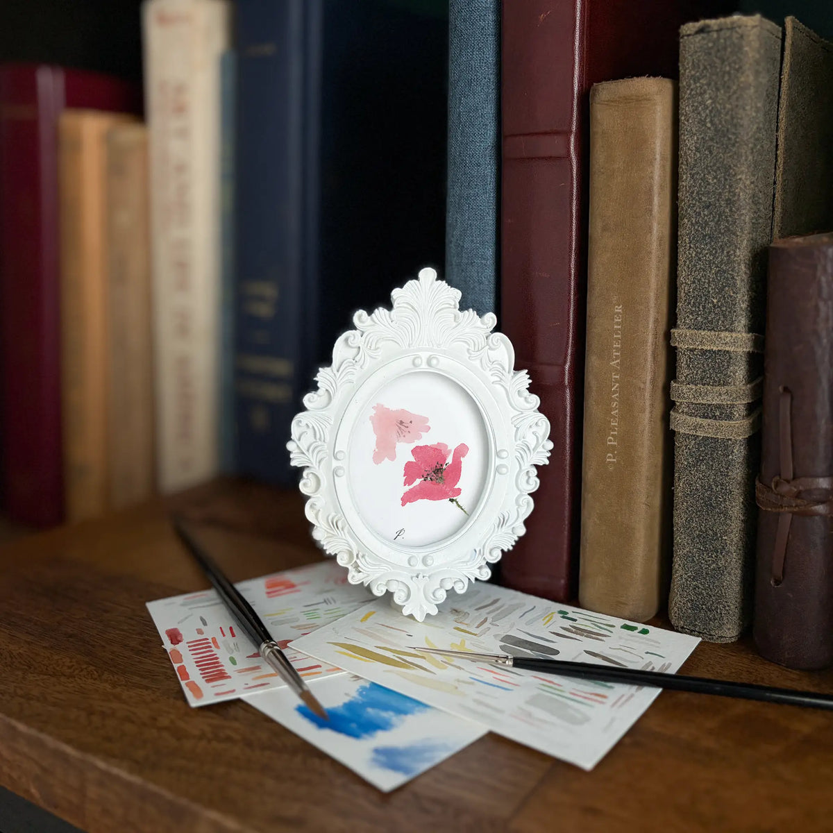 A small white ornate frame holding a pink flower print, styled on a wooden table with paintbrushes and art cards in front of a row of vintage books.