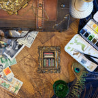 A styled flat-lay featuring a miniature gold-framed bookshelf holding colorful vintage books, surrounded by postage stamps, a peacock feather, a green glass vase, and a watercolor palette on a rustic wooden surface.