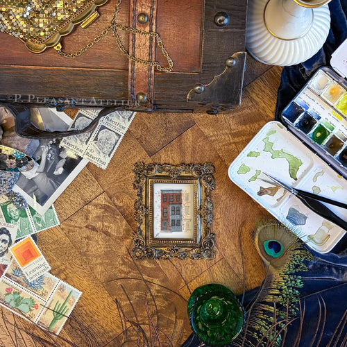 A styled flat-lay of vintage art supplies and framed prints on a wooden table. The central focus is a small ornate gold frame containing a painting of a red brick house, surrounded by a watercolor palette, peacock feather, green glass, and other antique-style items.
