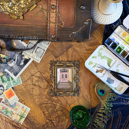 A styled flat-lay featuring a small framed art print of a pink door on steps, surrounded by vintage-style props including stamps, a watercolor palette, peacock feathers, and a wooden trunk on a wood floor.