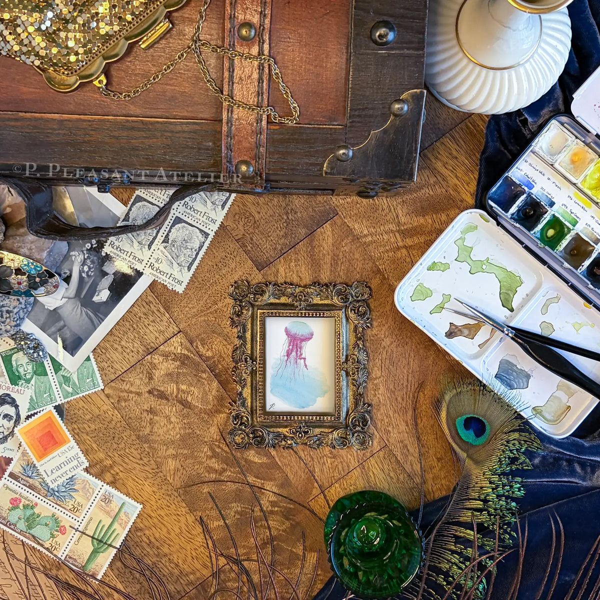 A styled flat-lay of vintage art supplies on a wooden table, featuring a small framed pastel painting of a jellyfish, watercolor paints, stamps, a peacock feather, and a wooden chest.