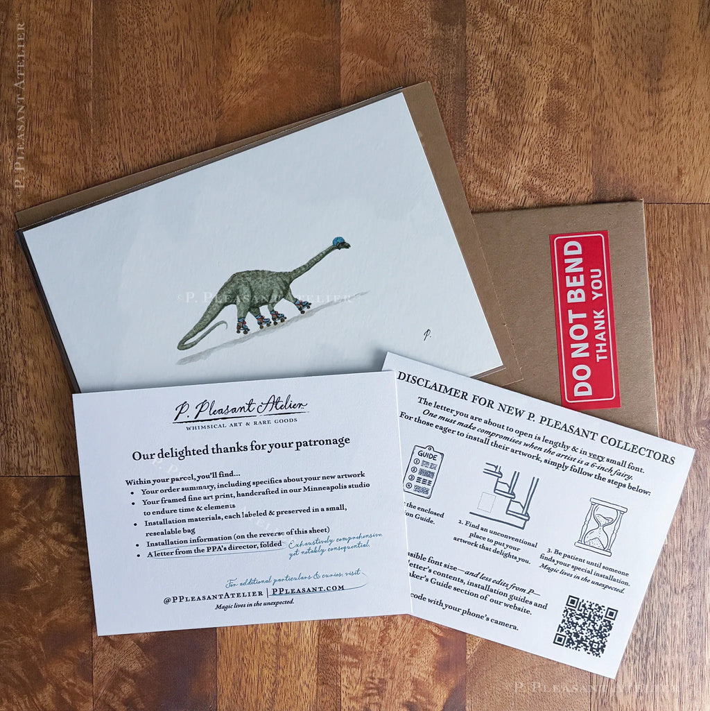 Example of the unframed artwork package laid on a wooden surface, showing a white card with a green dinosaur illustration, a brown envelope, a red 'DO NOT BEND, THANK' label, and two printed paperwork cards.