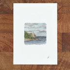 A vertical art print with a wide white border featuring a small watercolor painting of a coastal cliff and lighthouse, displayed flat on a brown wood plank surface.