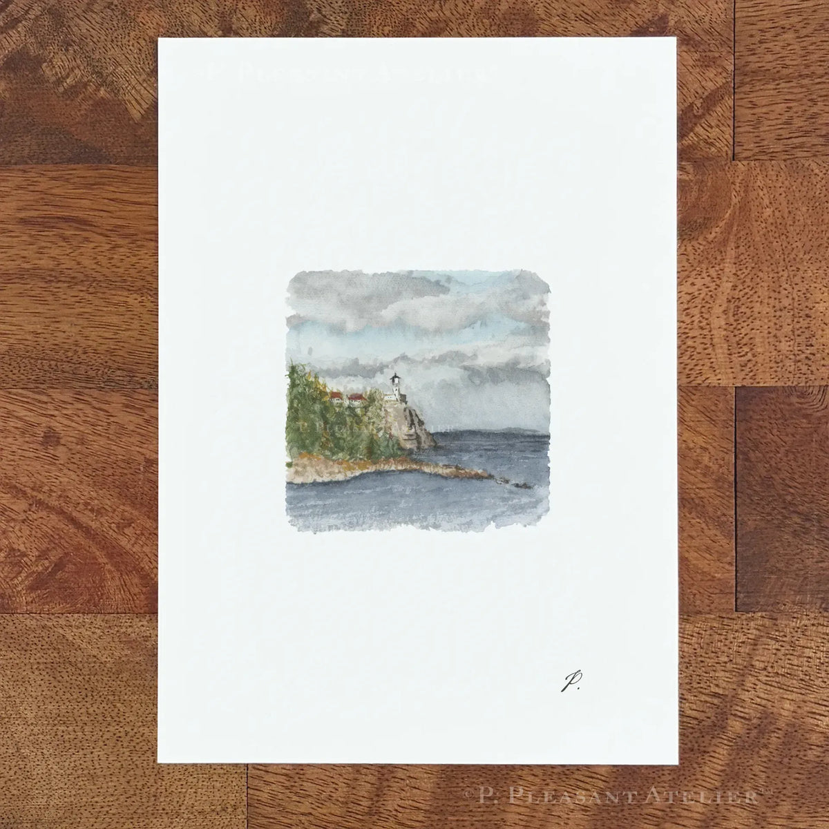 A vertical art print with a wide white border featuring a small watercolor painting of a coastal cliff and lighthouse, displayed flat on a brown wood plank surface.