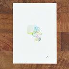 A vertical watercolor art print featuring a cluster of translucent bubbles in pastel shades of blue, green, and yellow on a white background, displayed flat on a brown wooden surface.